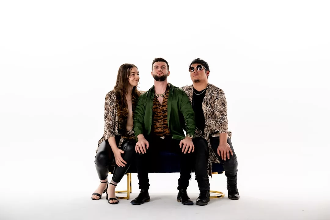 Three people sit on a bench against a white background; the man in the center wears a green jacket, while the two on either side showcase their fashion sense with animal print tops and dark pants.