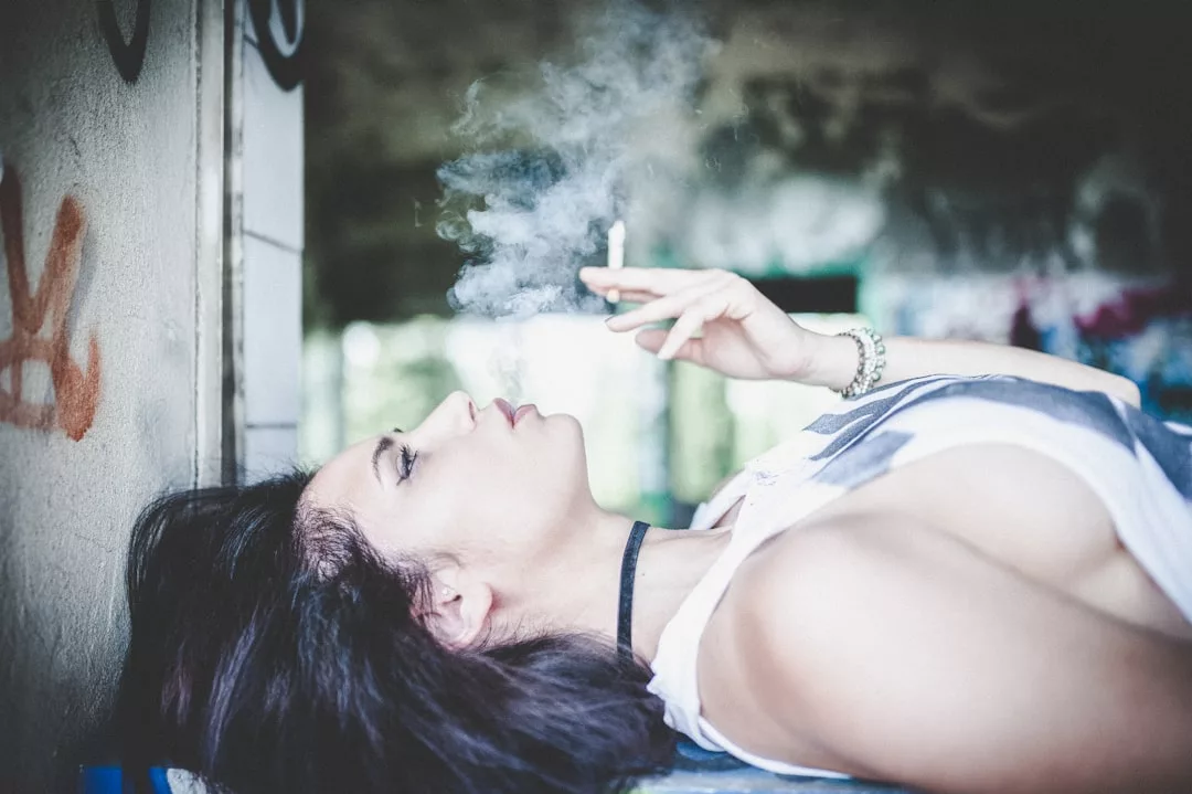 A person with dark hair lies on their back indoors, holding a lit cigarette and exhaling smoke. The scene appears in soft focus, with graffiti visible on the walls, capturing an edgy sense of fashion amid urban luxury.