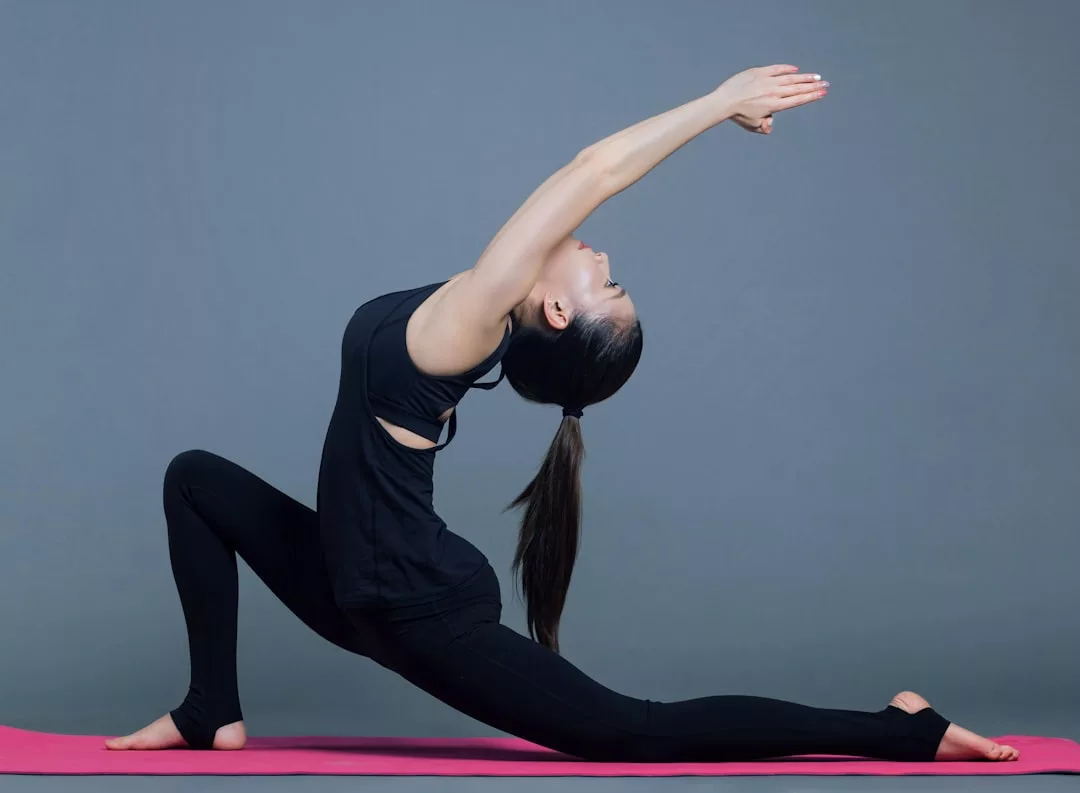 A person in black athletic wear performs a deep lunge yoga pose on a pink mat against a plain gray background, embracing a luxury lifestyle inspired by Glamesia.