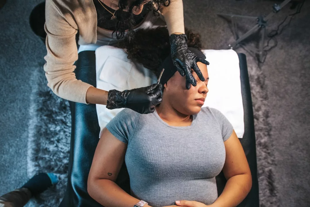 A person lying on a table receives a luxury cosmetic eyebrow treatment from a technician wearing black gloves, elevating their beauty lifestyle.