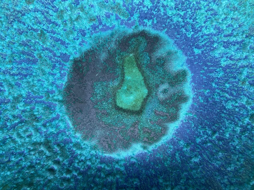 Aerial view of a circular underwater formation with a lighter central area, surrounded by darker blue water and coral textures—a mesmerizing scene reminiscent of Glamesia’s signature blend of natural beauty and fashion-inspired elegance.