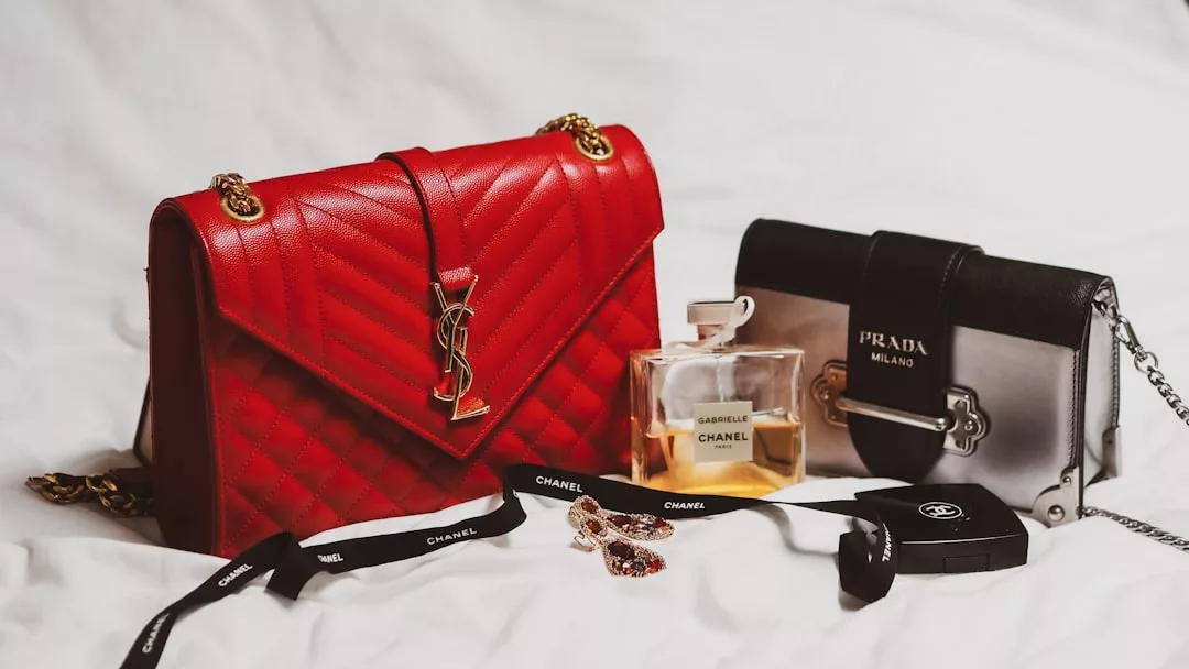 Luxury abounds with a red Yves Saint Laurent bag, black and white Prada bag, Chanel Gabrielle perfume, Chanel ribbon, sunglasses, and jewelry arranged elegantly on a white surface.