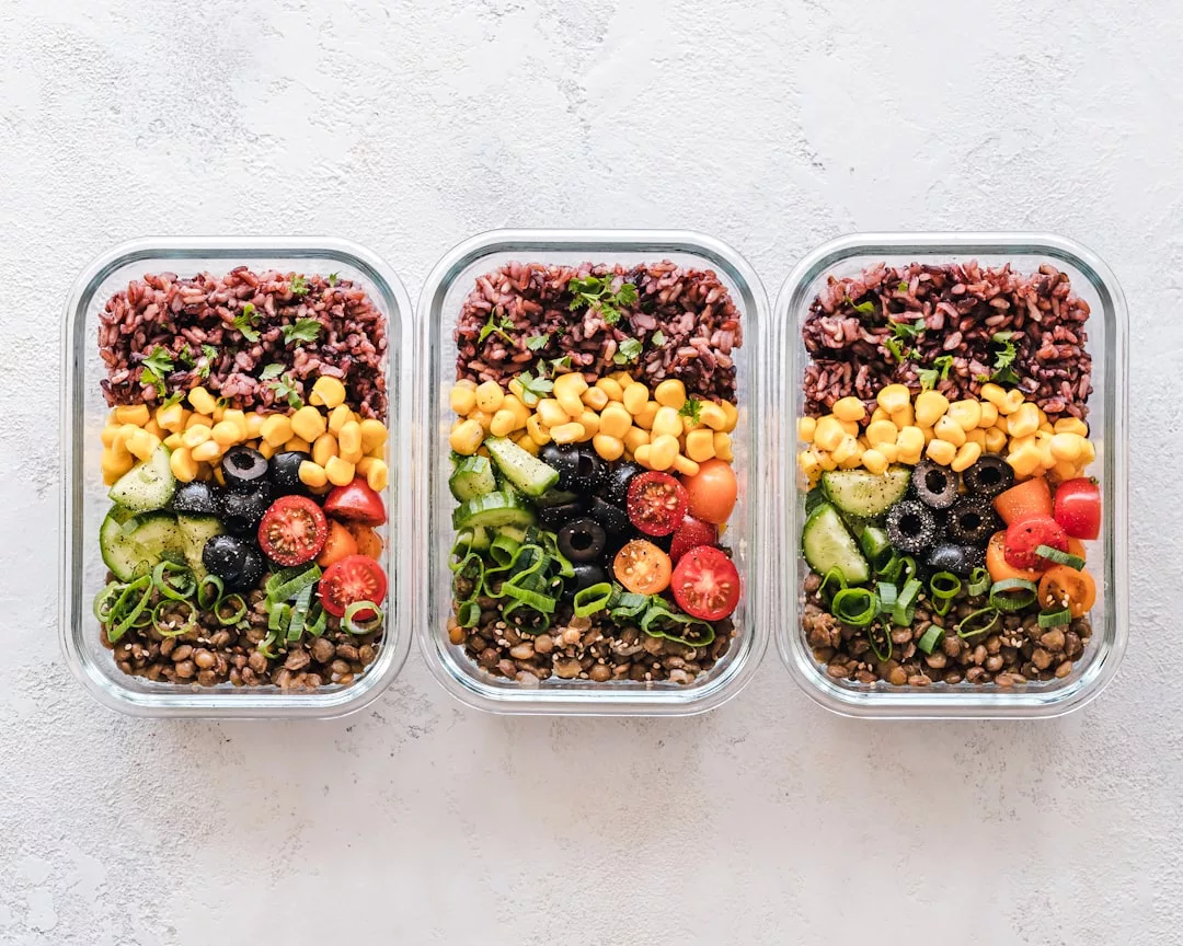 Three glass containers filled with brown rice, lentils, corn, cucumber, cherry tomatoes, black olives, and chopped green onions are arranged in neat rows on a light surface—adding a touch of luxury to your healthy lifestyle.