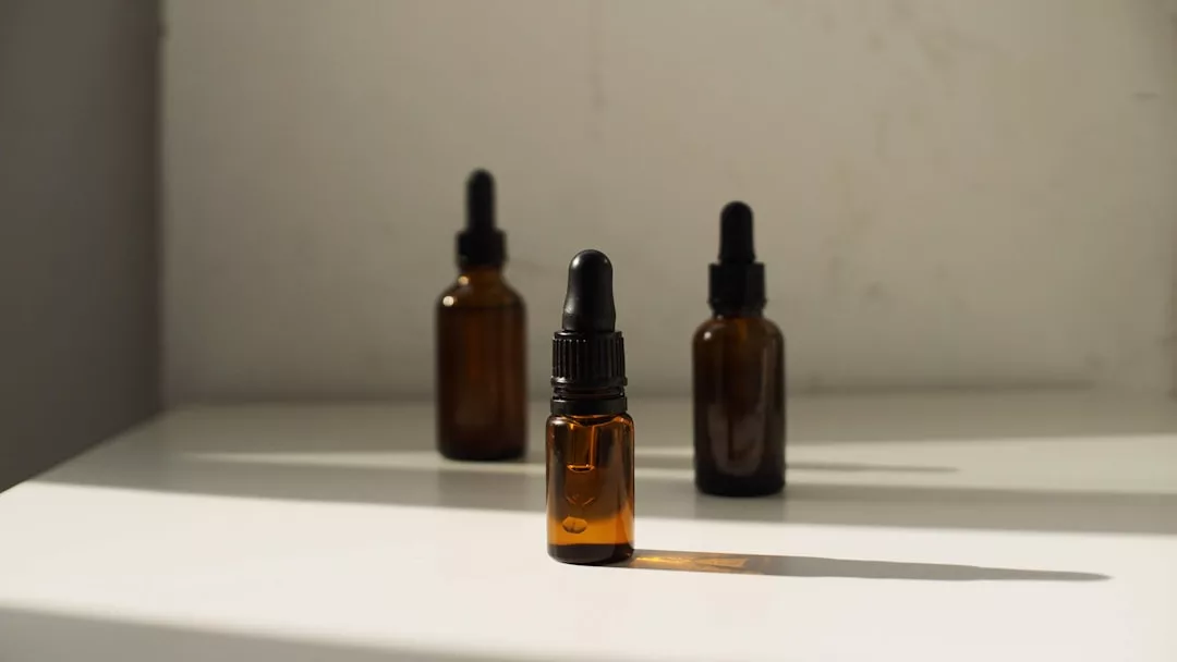 Three amber glass dropper bottles of varying sizes are placed on a white surface, with sunlight casting shadows behind them, evoking Glamesia’s signature blend of luxury and fashion.