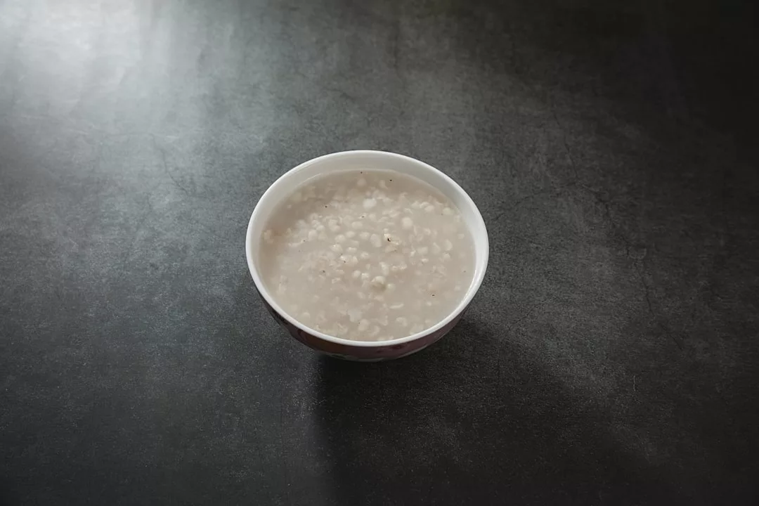 A white bowl filled with a light grayish, watery soup containing small grains sits on a dark, textured surface, evoking a sense of minimalist luxury in everyday lifestyle.