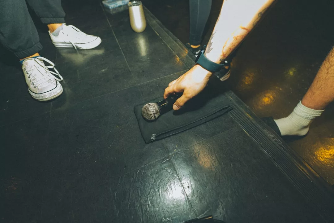 A person reaches down to pick up a luxury microphone lying on a black pouch on a dark floor, while others stand nearby.