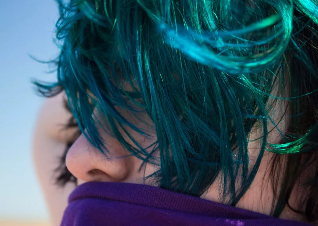 Close-up of a person with teal hair partially covering their face, wearing a purple garment up to their nose and eyes closed—a bold Glamesia fashion moment.