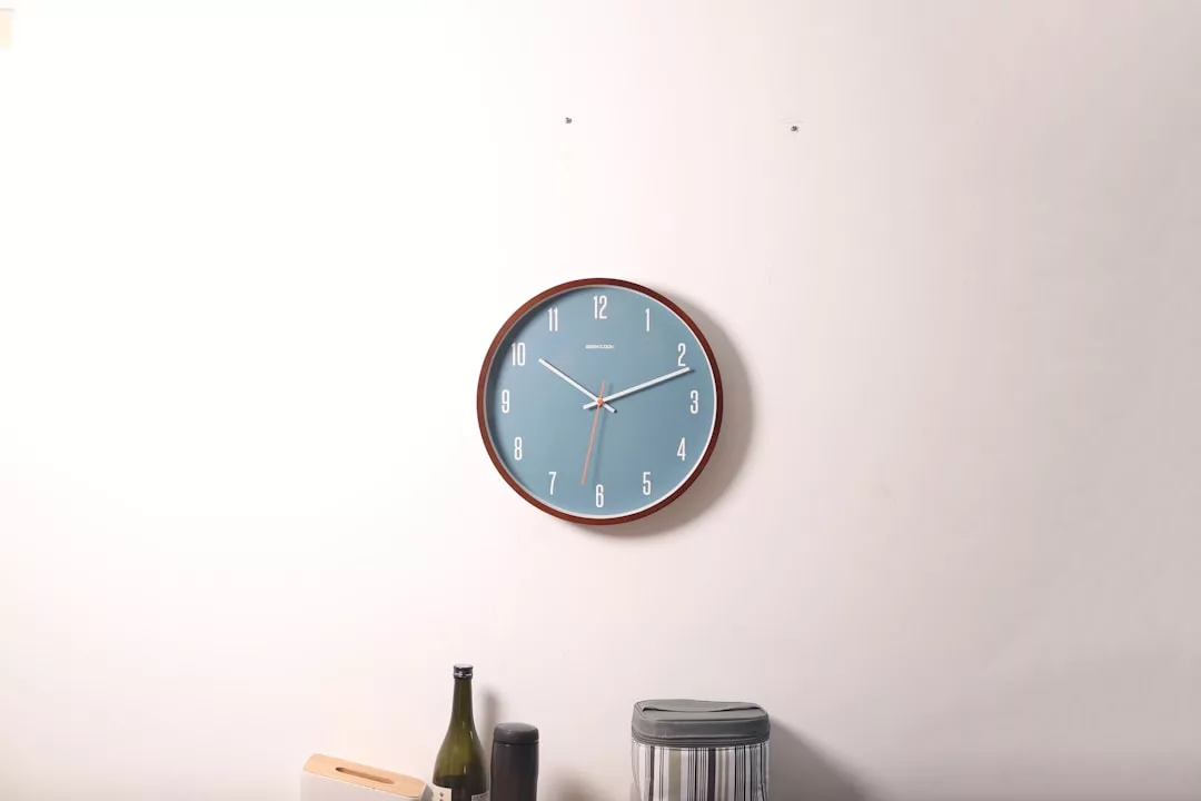 A blue Glamesia wall clock showing 4:40 is mounted on a plain white wall above a countertop with a wine bottle, a container, and a kitchen utensil holder, adding a touch of luxury to the setting.