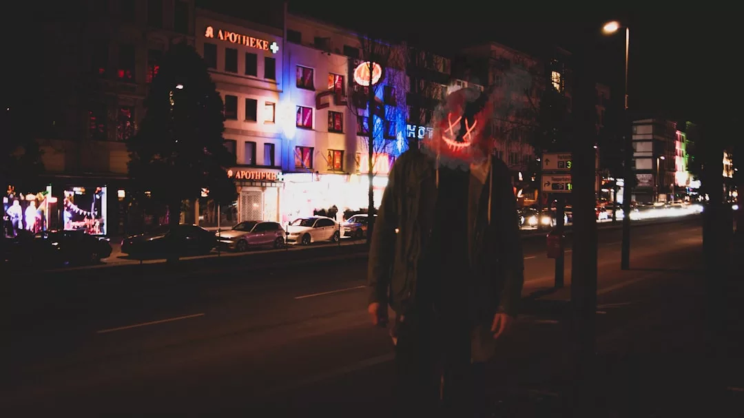 A person with a glowing mask stands on a dimly lit city street at night, exuding modern fashion amidst illuminated buildings and cars in the background.
