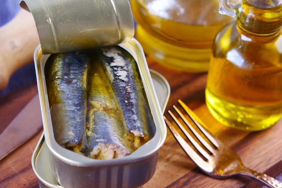 An open tin of sardines in oil sits on a wooden surface next to a fork, a glass bottle of oil, and a knife—perfect for enjoying a touch of luxury in your everyday lifestyle.