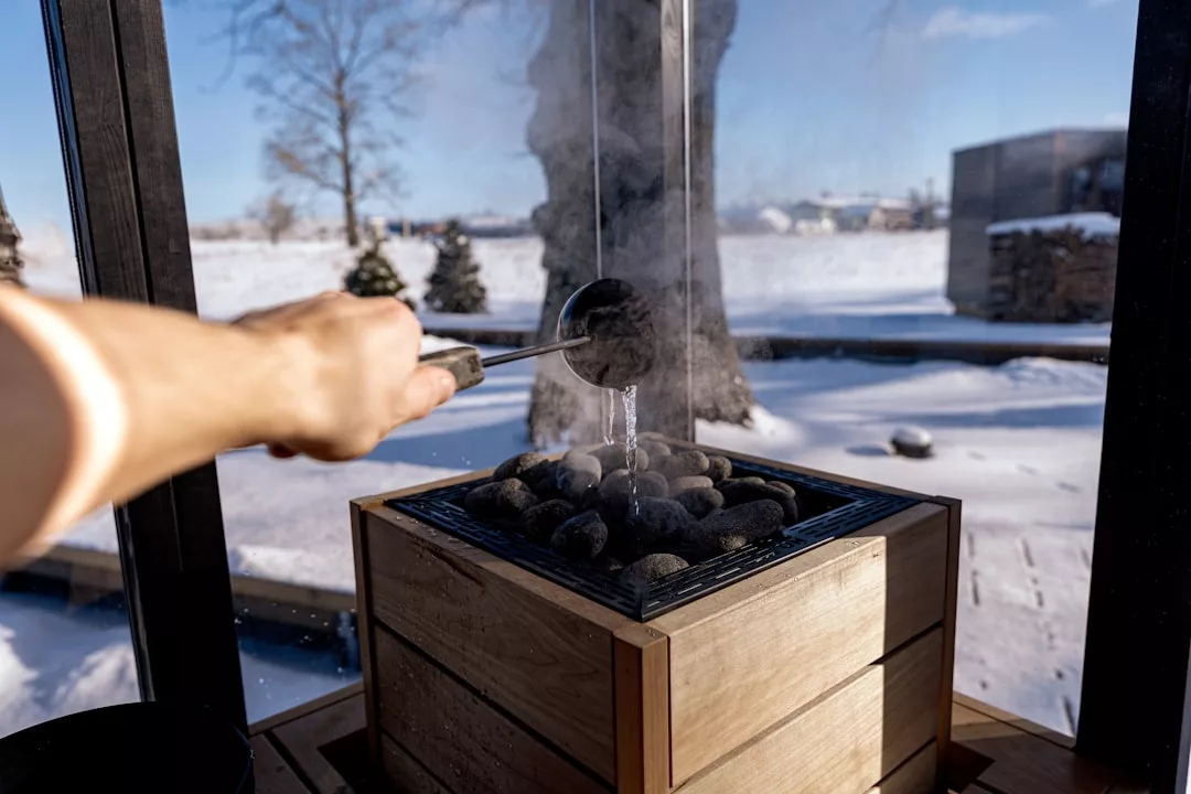 A hand pours water onto hot stones in a wooden luxury sauna stove, producing steam; a snowy outdoor scene is visible through the windows.