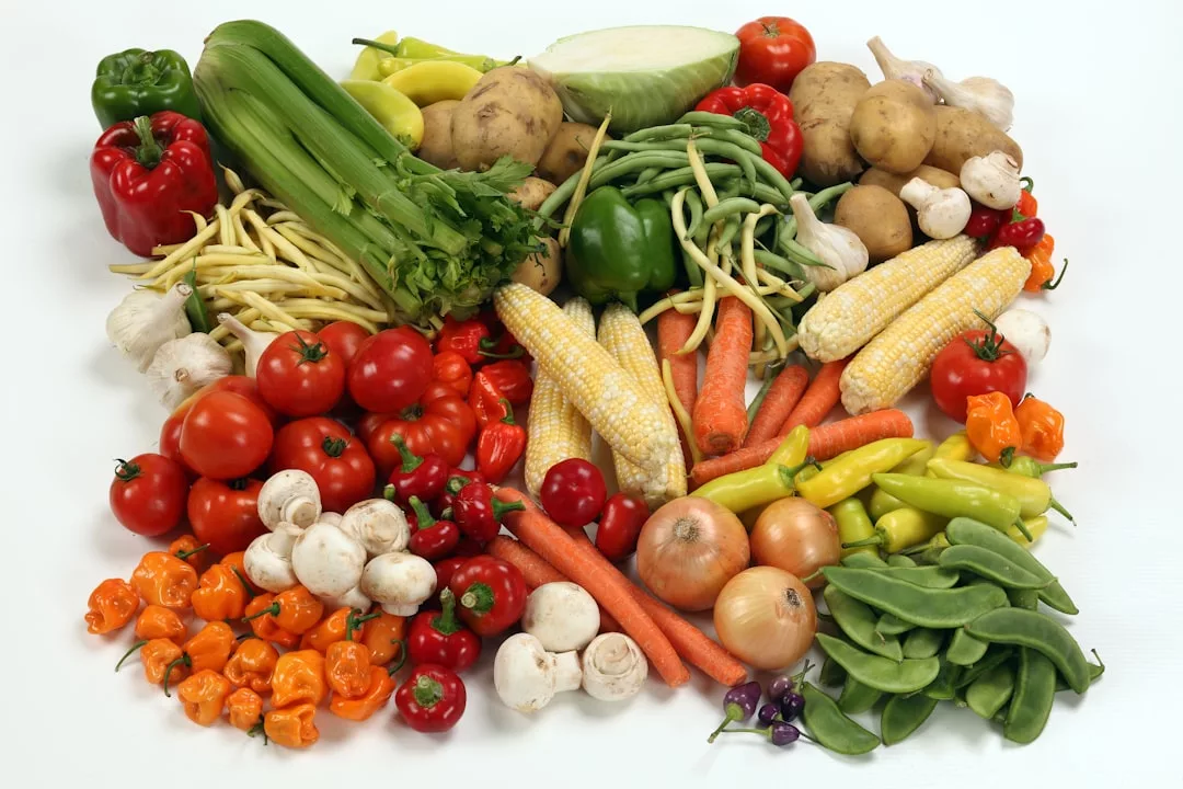 Assorted fresh vegetables including tomatoes, carrots, corn, onions, peppers, mushrooms, celery, beans, peas, potatoes, and cabbage arranged on a white surface evoke a Laxary touch to everyday freshness.