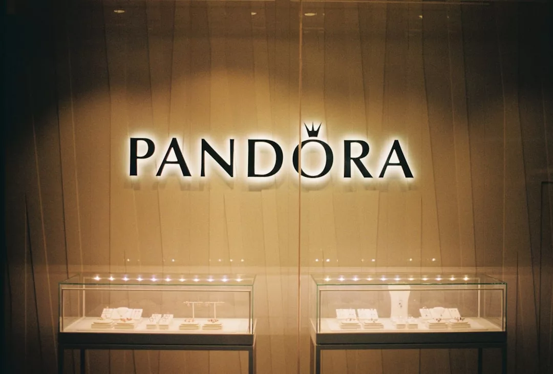 A Pandora store display with an illuminated Pandora logo above two glass cases, showcasing fashionable jewelry on a beige background for the modern lifestyle.