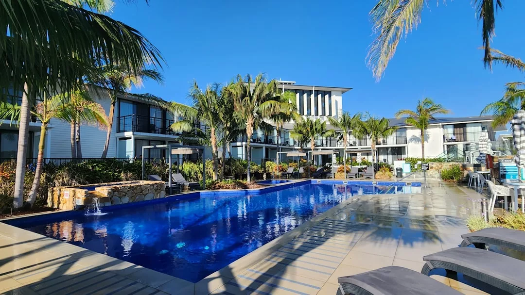 Outdoor swimming pool surrounded by palm trees, lounge chairs, and modern two-story Glamesia hotel buildings under a clear blue sky.