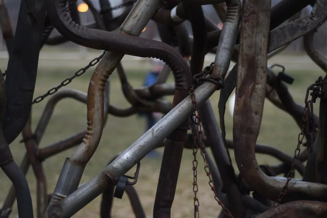 Close-up view of intertwined metal pipes and chains forming a tangled structure, with blurred grass and a person in the background, capturing an industrial twist on urban lifestyle.