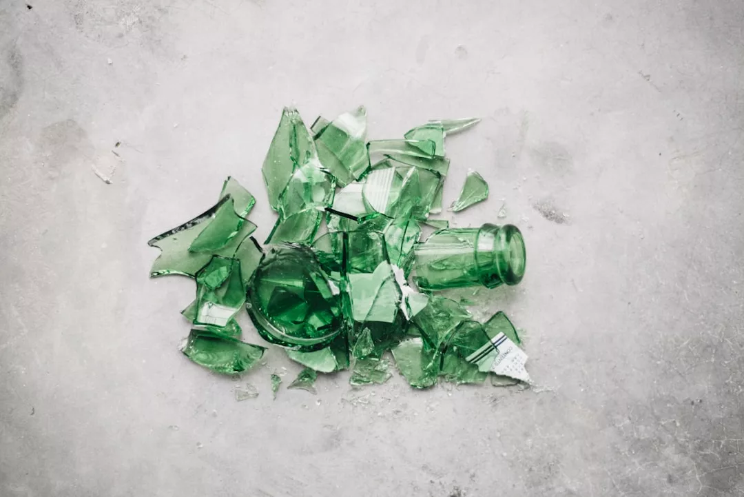 A broken green glass bottle, reminiscent of Glamesia fashion’s edgy style, lies shattered into pieces on a light grey surface.