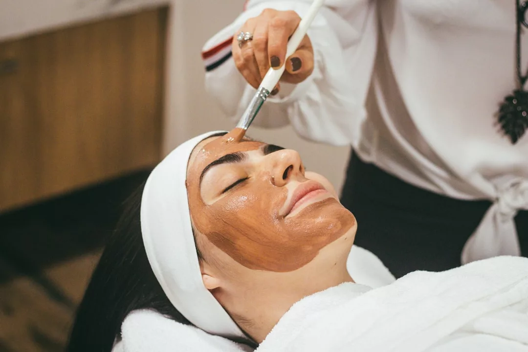 A person with a headband lies on a spa bed while another applies a brown facial mask with a brush, creating a Glamesia moment of luxury and relaxation.