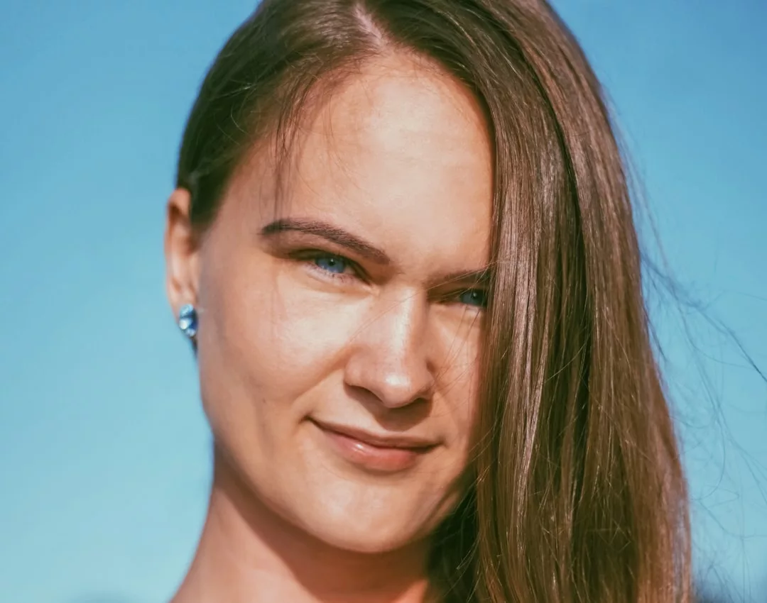A woman with long brown hair and blue eyes looks toward the camera, standing outdoors beneath a clear blue sky, radiating effortless fashion and luxury.