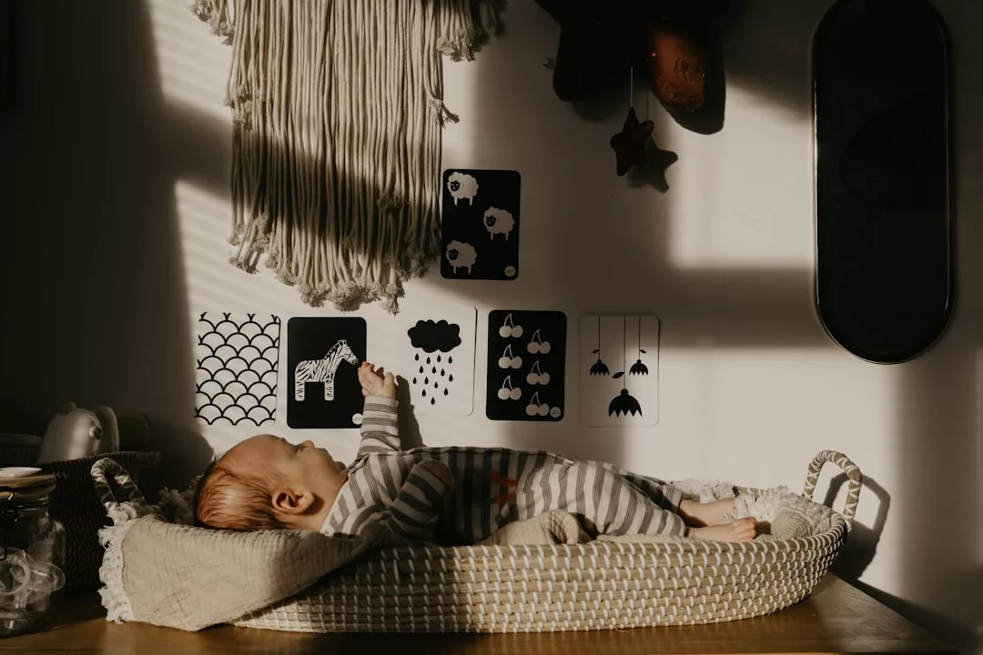 A baby in striped pajamas lies in a wicker bassinet, reaching toward black and white wall art in a softly sunlit room, embodying effortless Glamesia fashion.