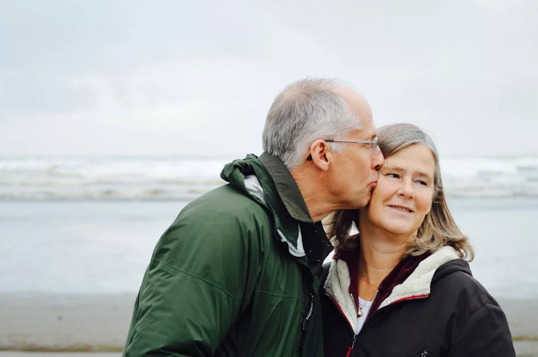 An older man in a green jacket shares a kiss on the cheek with a woman as they stand together on a beach, their moment framed by ocean waves and effortless fashion.