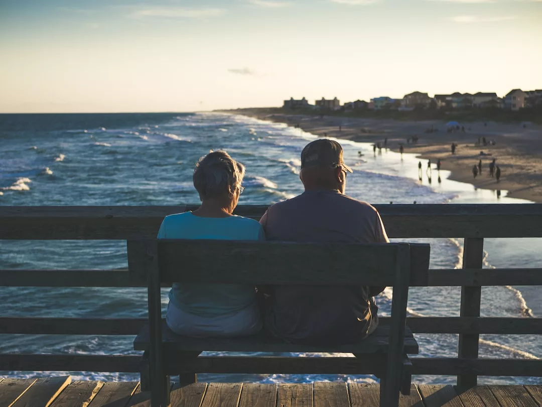 An elderly couple sits on a bench overlooking the ocean and beach during sunset, enjoying the gentle waves and people along the shoreline, embracing a moment of simple laxary amid nature’s beauty.