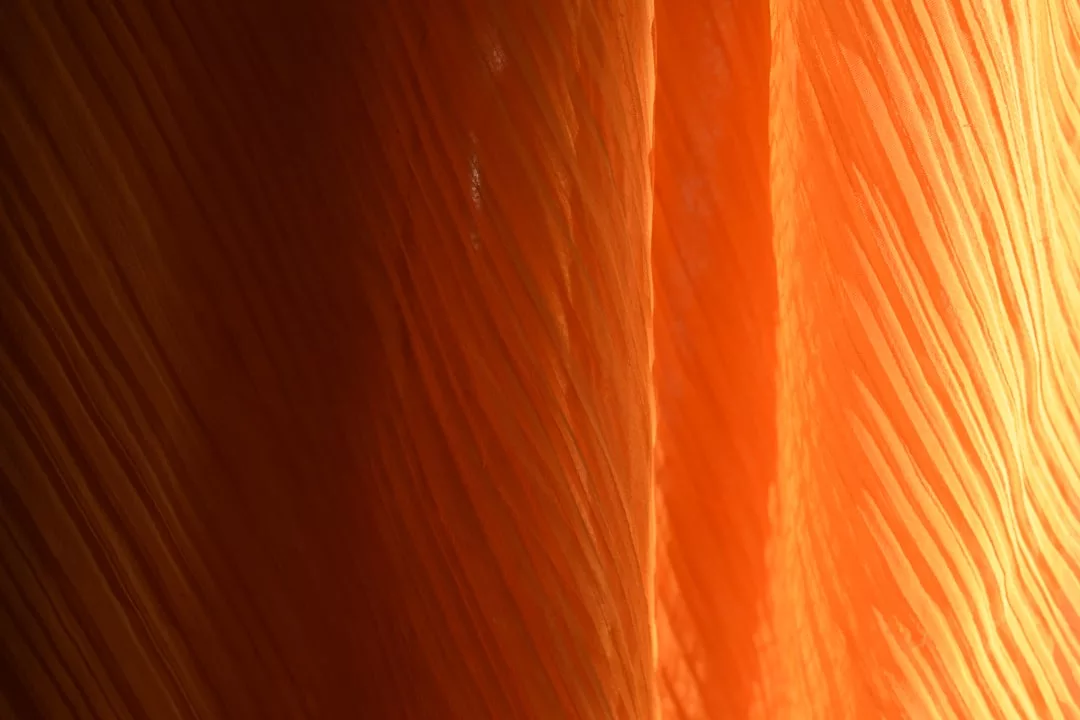 Close-up of orange pleated fabric with light shining through, highlighting the texture and vertical lines—a touch of luxury perfect for Glamesia fashion inspirations.