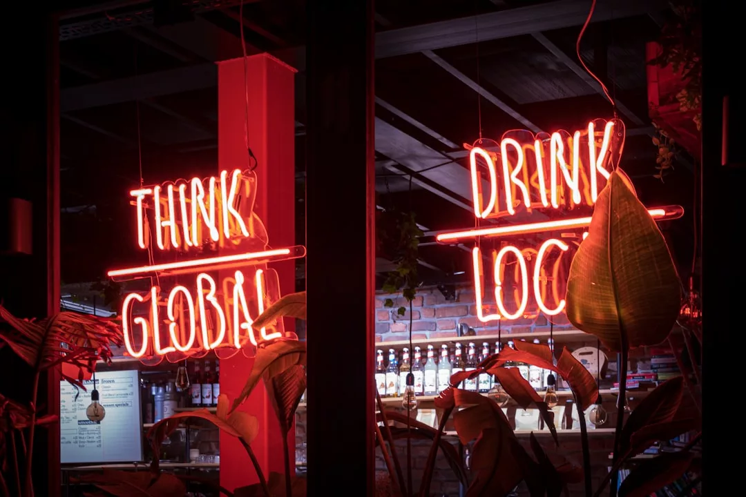 Neon signs in a bar window display the phrases "THINK GLOBAL" and "DRINK LOCAL," capturing a stylish lifestyle vibe, with plant leaves partially visible in the foreground.