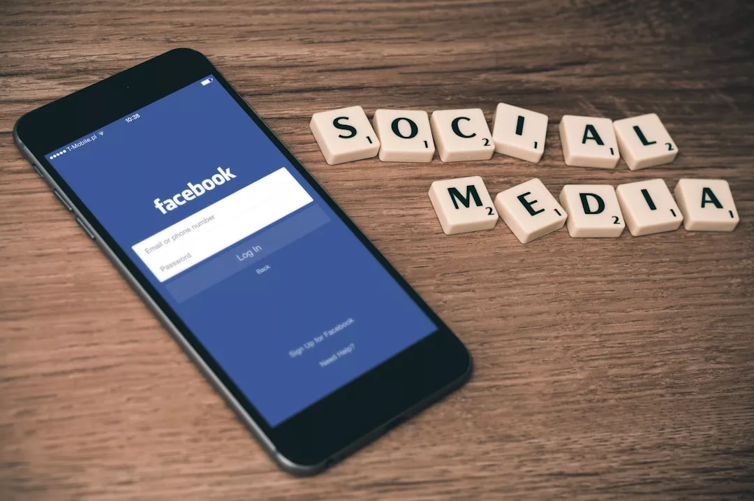 A smartphone displaying the Facebook login screen rests on a wooden surface next to Scrabble tiles spelling "SOCIAL MEDIA," creating a scene that fuses digital connection with luxury fashion vibes.