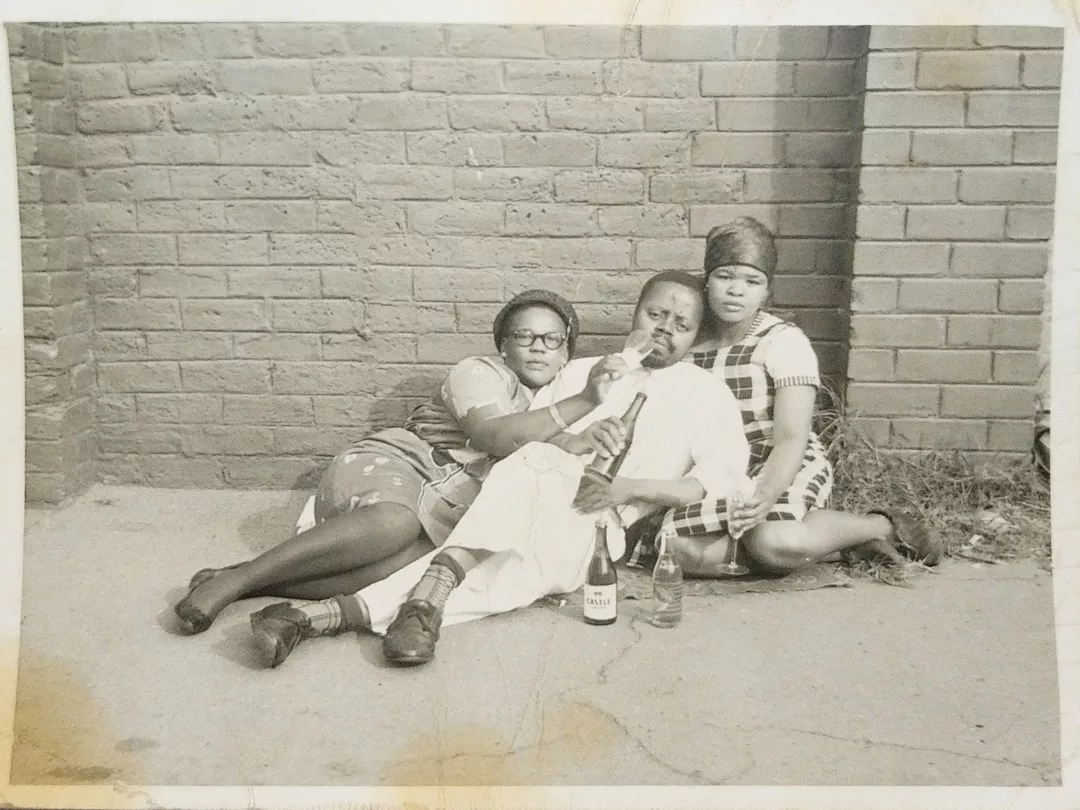 Three people sit on the ground against a brick wall, with two liquor bottles in front of them; their relaxed poses and stylish outfits add a touch of fashion as they all look directly at the camera.