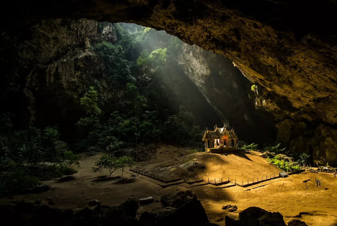 A small ornate pavilion exudes luxury as it sits illuminated by sunlight inside a large, dark cave, with trees and rocky terrain surrounding this Glamesia sanctuary.