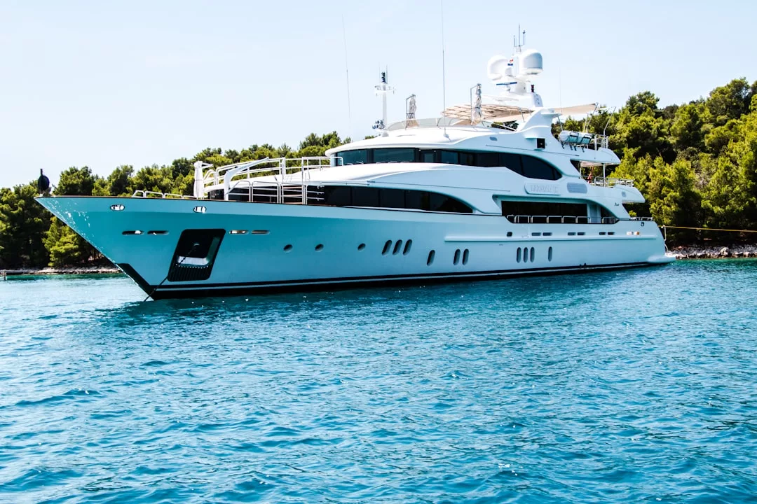 A large white luxury yacht is anchored on calm blue water near a tree-lined shoreline under a clear sky, exuding style and fashion.